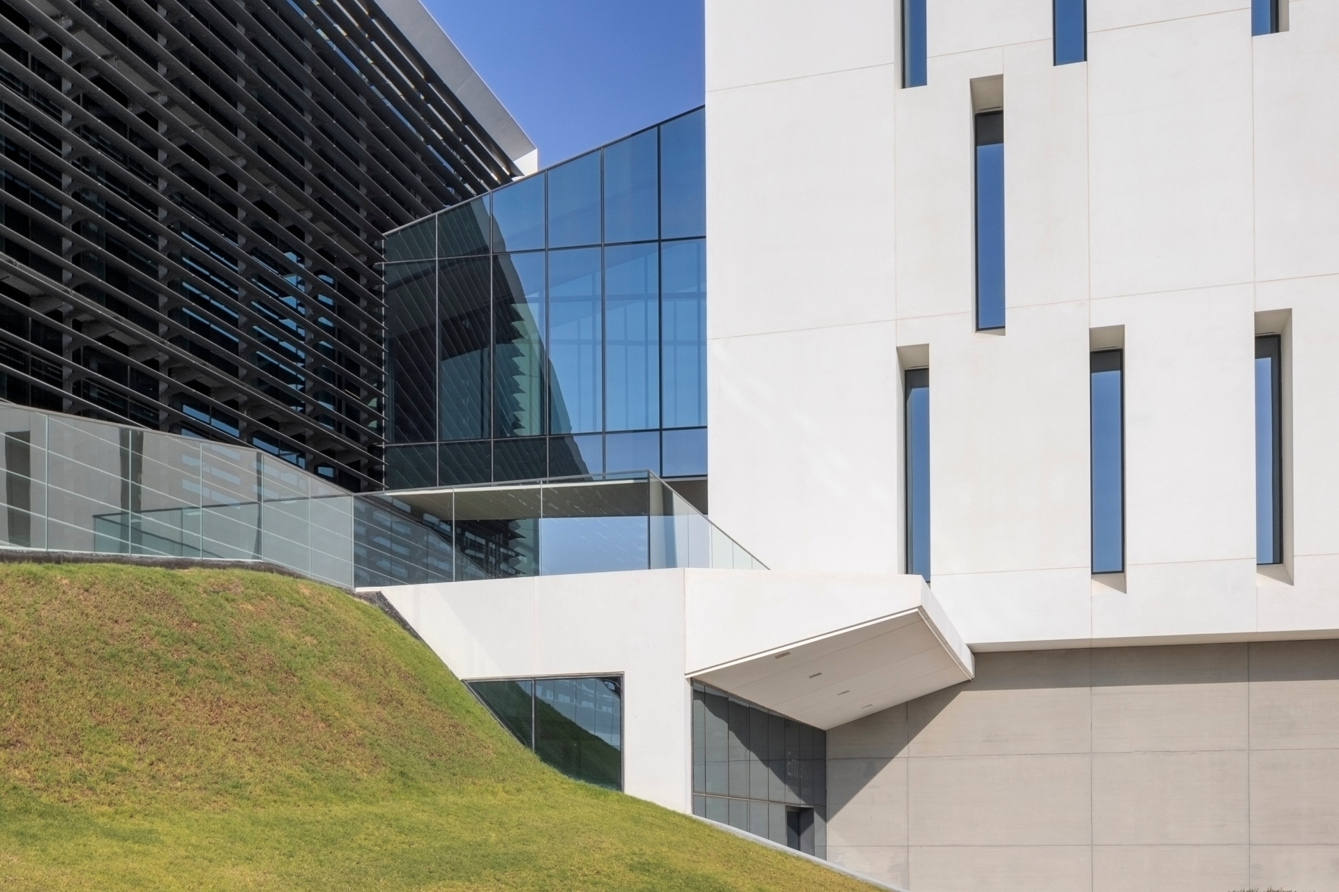 Jotun HQ — glass skywalk bridge connecting the dark louvred block and the white stone tower, with the landscaped grass berm and glass balustrade in the foreground