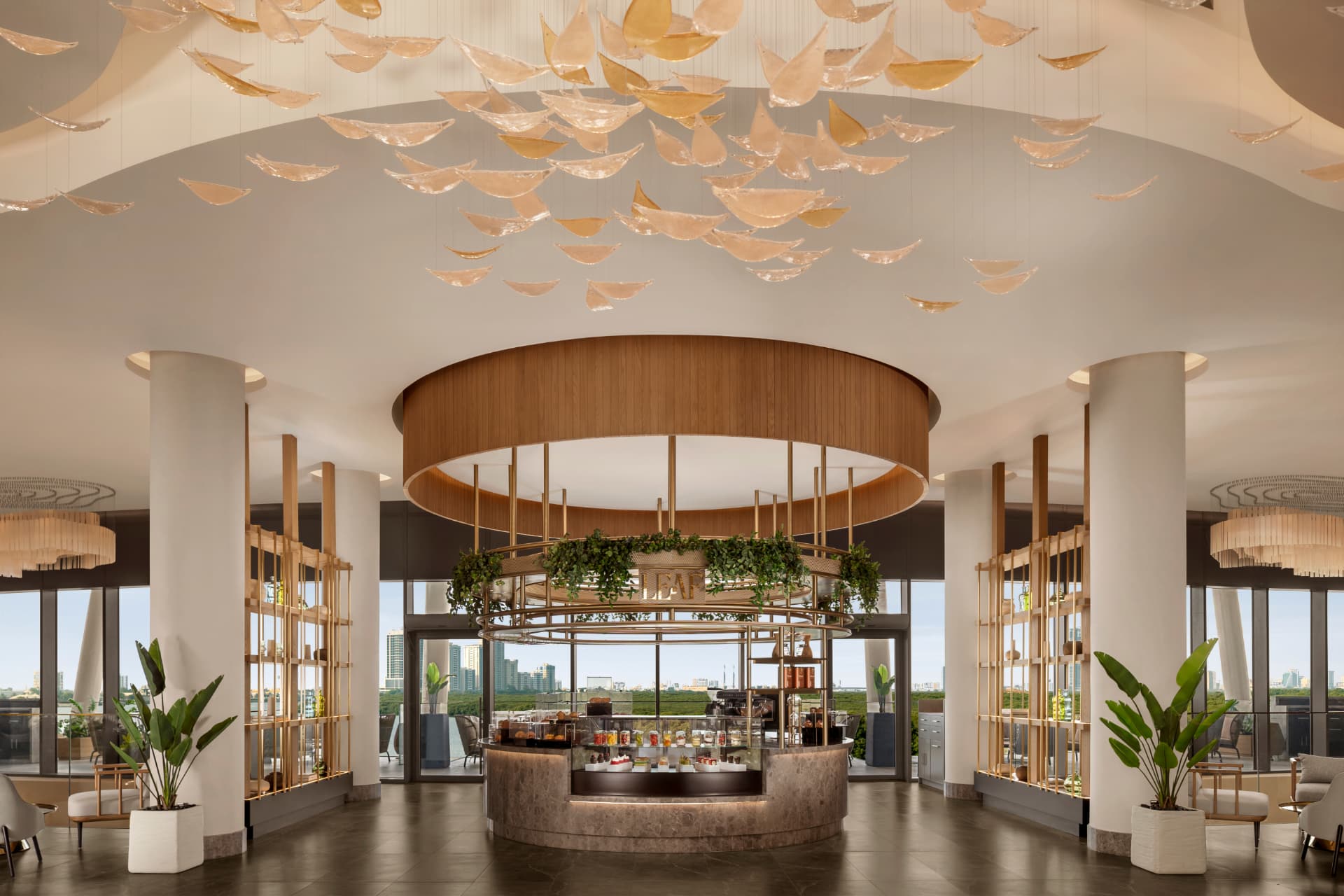 Circular lobby café at Conrad Dubai beneath a ceiling installation of golden leaf-like pendants, with city views through full-height glass