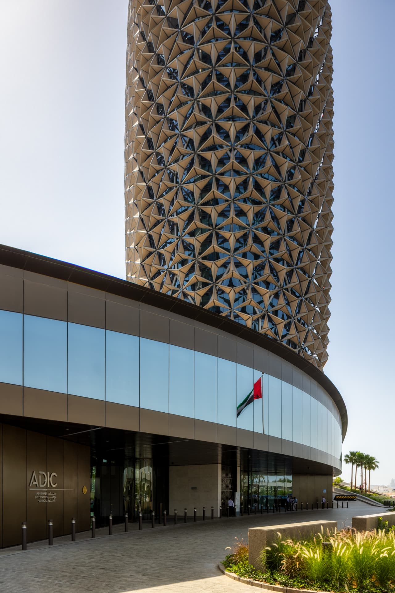 The curved base of the ADIC Headquarters tower meeting its glass podium entrance with ADIC signage, flagpole and palm trees