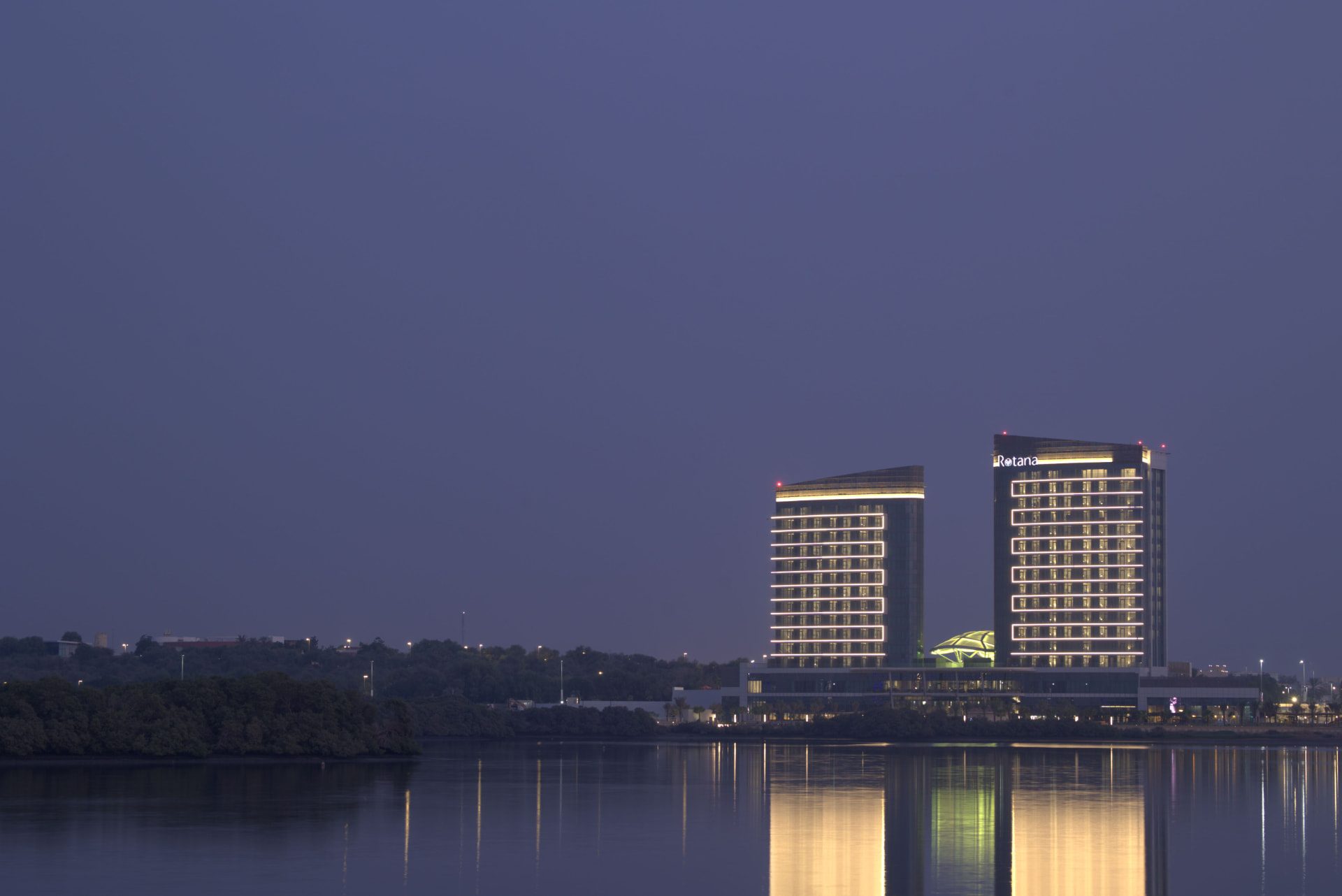 Rotana Ras Al Khaimah exterior before the refresh — flat, hazy dusk frame of the two illuminated towers across the mangrove channel with muted sky and weak reflections