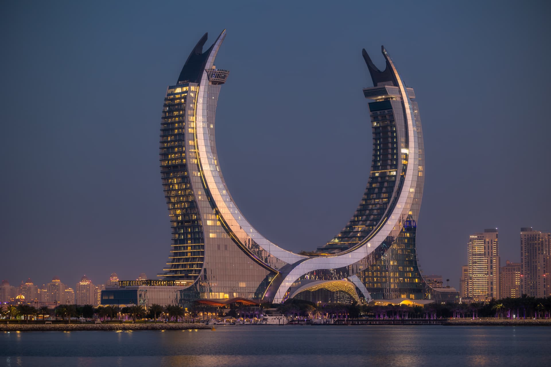 Crescent-shaped Katara Towers in Lusail at dusk, illuminated facades reflected in the bay beneath a deep blue sky